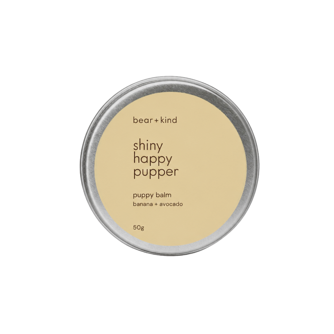 A round silver metal tin of bear + kind 'shiny happy pupper' puppy balm in banana + avocado scent, featuring a light beige label on a solid white background.